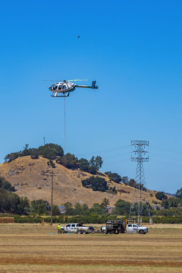 hero-img-01 Helicopter flying over rural landscape with vehicles in grass field under clear blue sky.